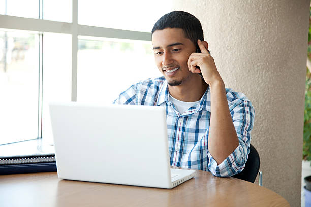 A Hispanic student surfing the web while making a call Latin student smiling while looking at his white laptop  and is talking on his iphone on a college campus. puerto rican ethnicity stock pictures, royalty-free photos & images