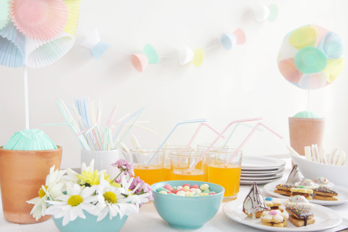 A DSLR photo of a table setting decorated for a birthday child party or baby shower. Decoration with two cupcake liner topiary at the sides and cupcake liner garland on the background. A blue vase with white daisies are in the foreground at the left. Drinks with orange lemonade are served in the middle of the table. Plates with sandwiches, sweet pastries and candies sits on the foreground at the right side. Main colours are white, pale pink, pale green, pale blue and yellow.