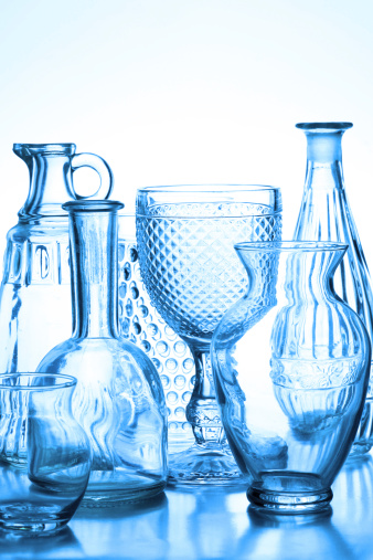 A studio shot of a collection of glass vases, wine glasses and glass bottles back lit toned in blue color in a reflective surface. Glasses features a variation of finished surfaces like bubbles, ruffled and diamond pattern and a variation of shapes.