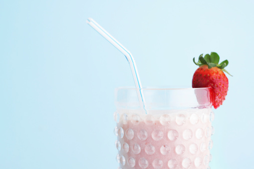A studio close up shot of a strawberry smoothie drink served with a drinking straw and decorated with a strawberry fruit. The smoothie is served in a hobnail clear glass cup. Pale blue background. Crop view of the top of the drink. Copy space.