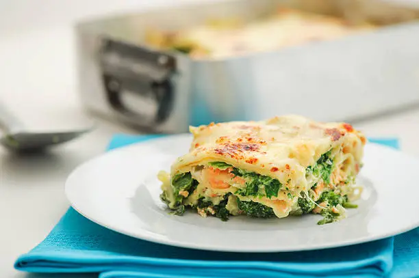 A plate with a nice piece of salmon lasagna with spinach leaves. Selective focus shallow depth of field on a clean, white background.