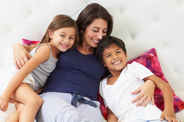 madre y niños relajante en la cama usando pijama - familia con dos hijos fotografías e imágenes de stock