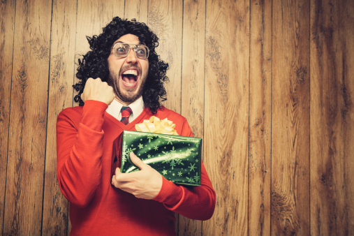 An ugly man with long curly black hair and 1980's retro style cheers with excitement in front of a wood paneled wall holding a Christmas present. He wears a Christmas colored red and green tie and sweater.  Horizontal with copy space.