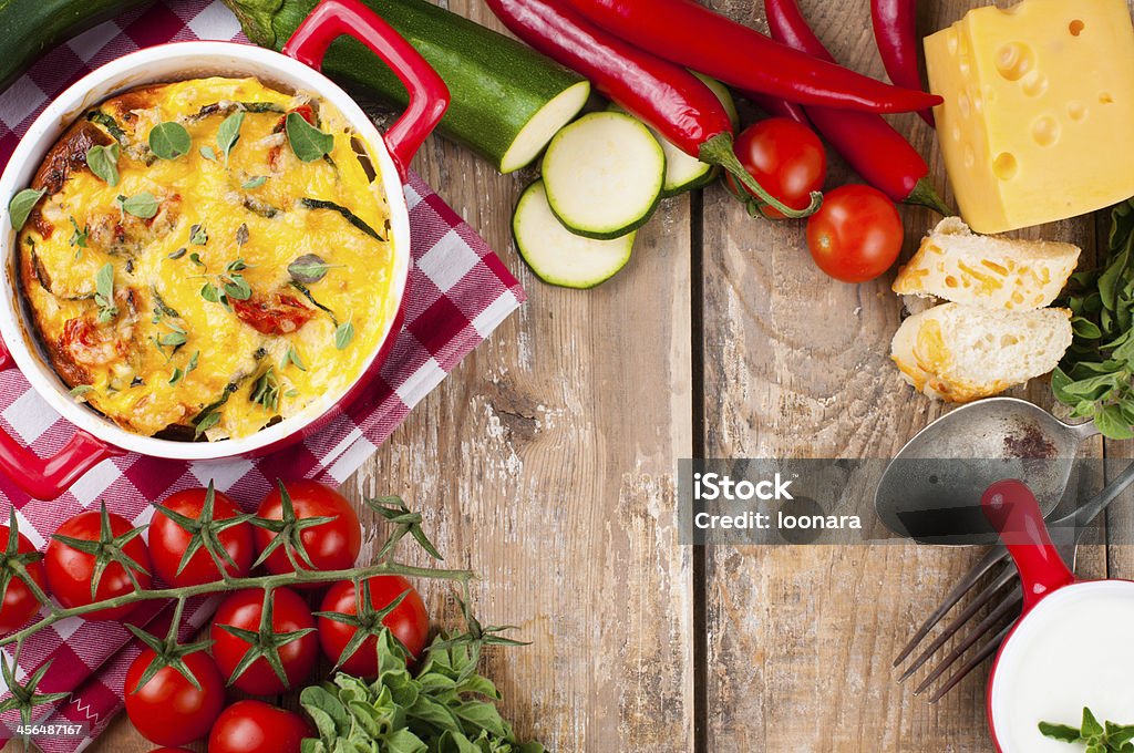 Vegetable casserole in a red pot Vegetable casserole in a red pot with cheese, zucchini, cherry tomatoes, oregano and cream sauce on a wooden board, home cooking Backgrounds Stock Photo