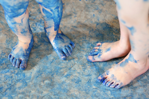 two pairs of very messy blue painted kid's feet are standing on a floor covered in paint