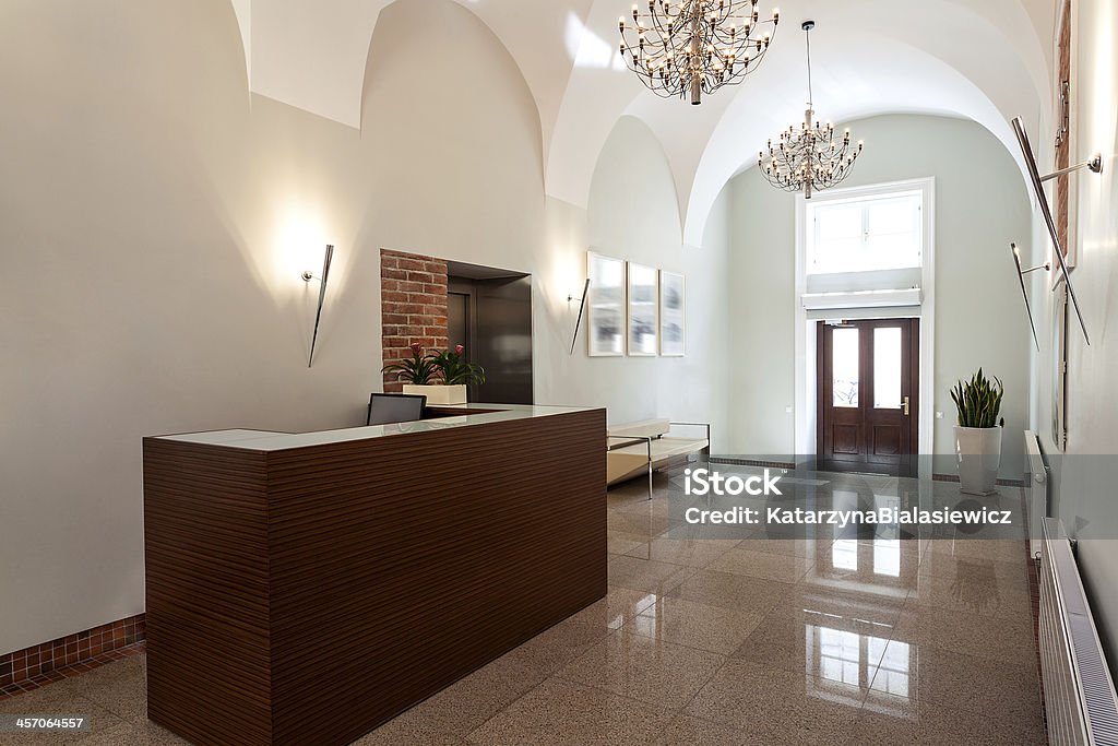 Entrance and reception area of elegant hotel This hotel reception area shows a brown reception counter, contrast against a white floor, off-white walls and high, arch ceilings.  The reception desk is in the forefront on the left side of the frame, with the main entrance door, with a brown wood frame and two, glass panels, is in the background.  Two chandeliers hang above the reception area. Apartment Stock Photo