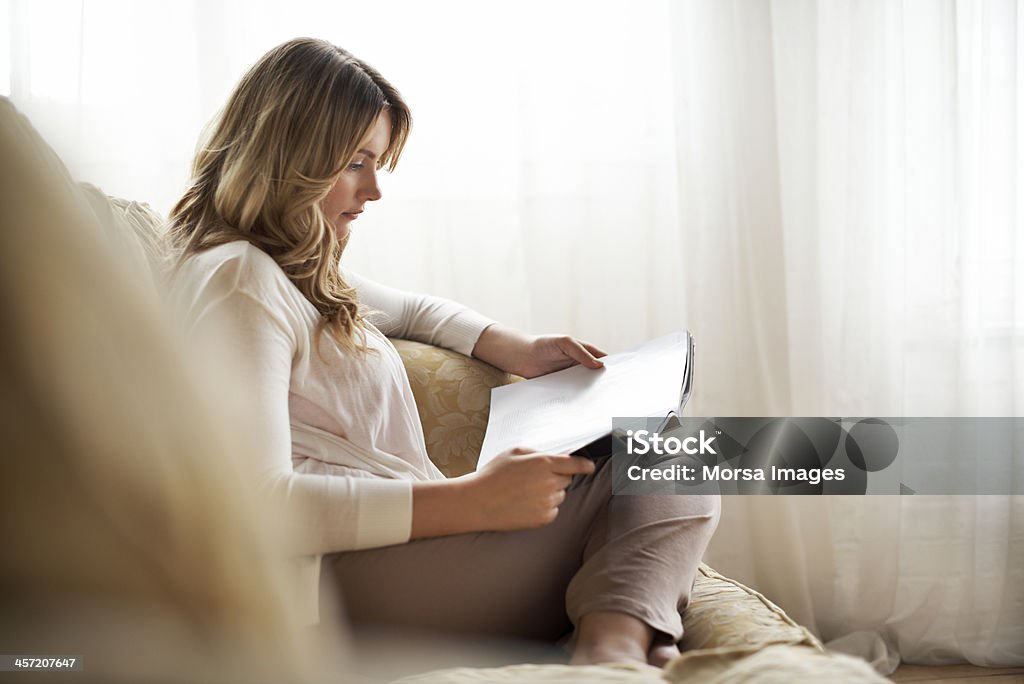 Woman sitting on sofa reading magazine Woman sitting on sofa reading magazine in her cozy loft apartment Reading Stock Photo