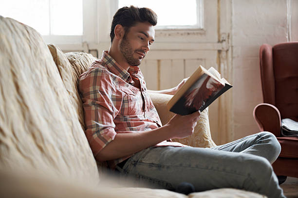 man sitting on sofa reading a book - reading book men sofa foto e immagini stock