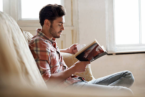 man sitting on sofa reading book - junger mann allein fotos stock-fotos und bilder