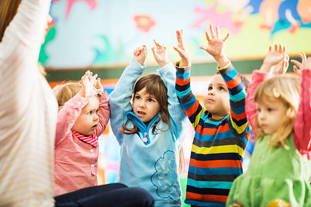 Kids playing with their teacher. Cute children playing in the playroom with their arms raised.   two groups stock pictures, royalty-free photos & images