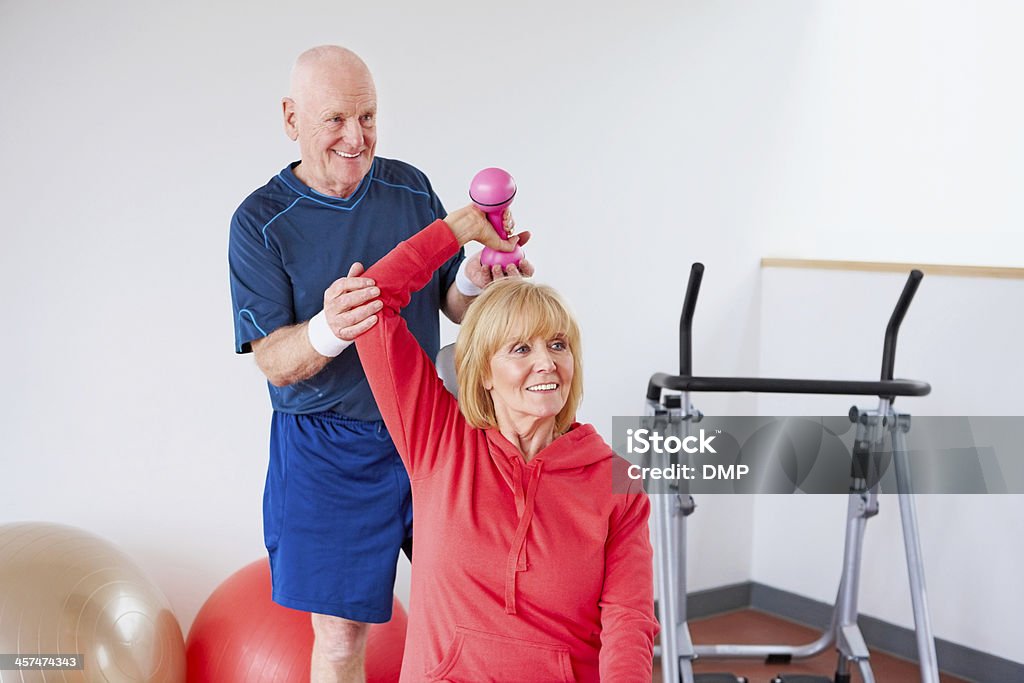 Active seniors exercising in gym together Senior man supporting woman exercising with dumbbell 60-69 Years Stock Photo