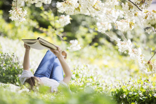 женщина чтение книги в траве под дерево с белыми цветами - book reading white women стоковые фото и изображения