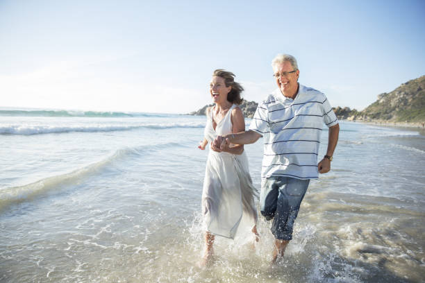 pareja de ancianos jugando en las olas en la playa - 60 65 años fotografías e imágenes de stock