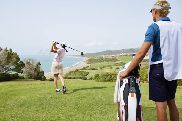 caddy watching woman tee off on golf course overlooking ocean - golf swing golf teeing off men стоковые фото и изображения