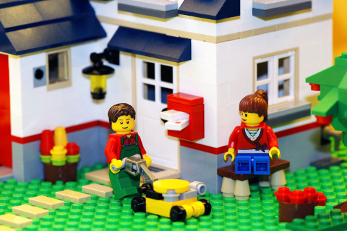 Borgosesia, Italy - October 10, 2011: Happy couple outside their new house built with the famous Lego construction block toys. Yellow light to simulate sunlight. The man is using a mower while the woman is sit on a bench.