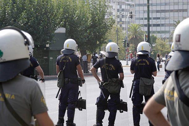 Behind Greek Riot Police Athens, Greece - September 8, 2011: Posterior view of Greek Riot Police waiting for rioters near Syntagma Square in Athens, Greece in the daytime.  The police are all men and are in a row, to create a human shield.  They have plexiglass shields and white helmets for their protection.  The word "POLICE" is written on the back of their uniforms in both English and Greek.  They also have batons and other equipment on their belts.  The posterior view shows what they are viewing on the street - there are no rioters yet, just pedestrians walking with city buildings in the background.  There has been violent rioting by protestors since the collapse of the Greek economy and the implementation of austerity measures on the Greek people. riot police stock pictures, royalty-free photos & images