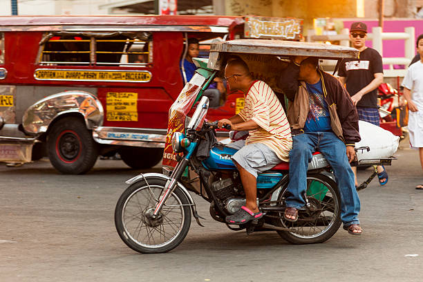 Driver and passenger on a motorcycle rickshaw, Metro Manila "Manila, Philippines - April 19, 2012: Driver and passenger on a motorcycle rickshaw known as a ""Tricycle"" in Metro Manila people and a Jeepney can be seen in the background." philippines tricycle stock pictures, royalty-free photos & images
