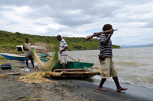 Lake Chamo, Ethiopia - August 8, 2010: In Nechisar National Park, not far from the town of Arba Minch, three Ethiopians stand on the lakeshore. Two are tending to a fishing net. One other is walking with a rifle on his shoulder, carried for self-defense given the history of tribal conflict in the area.