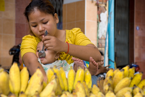 Phnom Penh, Cambodia - June 25, 2007: A Cambodian woman selling bananas at a street market feeds her baby with a spoon.