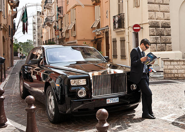 Chauffeur stands by Rolls Royce reading. "Monte Carlo, Monaco - May 2 2011. Chauffeur stands by black Rolls Royce car reading magazine, waiting in street of Monaco." rolls royce stock pictures, royalty-free photos & images