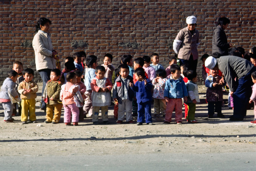 Beijing, China - Nov 8, 1986: Toddlers in colourful outfits are waiting by the road in the bright sunshine outside a Beijing nursery, while one of the four staff comforts a crying child. To avoid getting lost or straying onto the road, the children all have yellow rings tied to a string to hold on to.