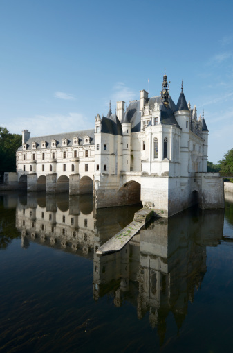 Horizontal landscape photo of the cobblestone paved walkway leading to the former drawbridge entrance in the surrounding wall of the castle at Saumur. Trees, Castle towers and rooftops can be seen in the distance under a blue sky. Saumur, Maine-et-Loire, France. 1st April, 2019