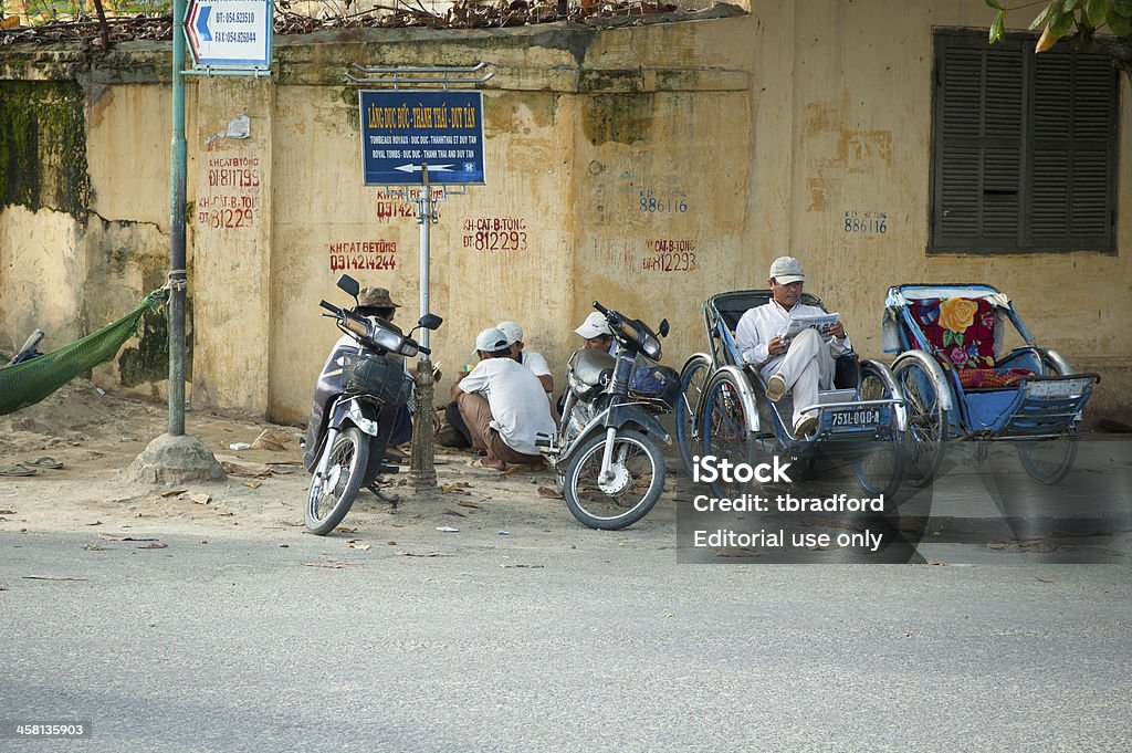 Motorcycle Taxi And Rickshaw Drivers Wait For Customers In Hue "Hue, Vietnam - December 8, 2006: Motorcycle taxi and rickshaw drivers playing card games and reading whilst waiting for customers. Motorcycle taxis and rickshaws are a popular form of public transport in Vietnam. The drivers have written their names and phone numbers on the wall." 2006 Stock Photo