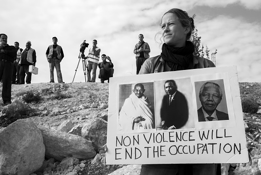 Bil'in, West Bank, Palestinian Territories - December 8, 2006: A European woman standing in a field near the Palestinian village of Bil'in, demonstrating against the Israeli occupation and separation barrier. In the background are media and a medic (holding first aid kit). The woman's placard features images of Gandhi, MLK Jr, and Nelson Mandela.