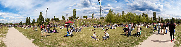 Mauerpark Flee Market Sunday Panorama "Berlin, Germany - June 10th, 2012: Wide angle 180 degrees Panoramic view of Mauerpark on an early summer Sunday, packed with people relaxing on the grass or engaged in various leisure activities. Sundays at Mauerpark are famous for their large flee market along with various shows held by all kinds of performers. The paths on the sides of the image are actually one and the same path." mauer park stock pictures, royalty-free photos & images