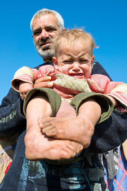 Atimah, Syria - January 14, 2013: A Syrian man holds a barefoot two-year-old child in Atimah refugee camp in northern Syria, home to some 12,000 people displaced by the country's civil war. These two had fled the city of Jisr al Shughour five months earlier and said they were struggling for basics, including shoes for the young children.