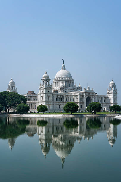 Victoria Memorial, Kolkata, India. Kolkata, India - October 04, 2011: As the name suggests the Victoria Memorial is a memorial to Queen Victoria. This photograph is taken slightly at a tangent from the main entrance which can be clearly seen in the photo. Furthermore the foreground is one of the several water features which figure around the building. The water feature reflects the front facade and three corners of the Memorial. The shot was taken late morning with a perfectly clear blue sky and very little wind. Furthermore, the shot is clear of people. The angle of the photograph show the varied influences in the architectural styles deployed to give it that uniqueness which even today the Victoria Memorial enjoys despite its colonial past. kolkata stock pictures, royalty-free photos & images
