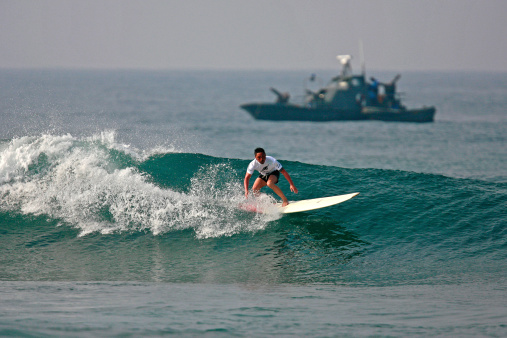 Hikkaduwa, Sri Lanka - March 21, 2010: Foreign Surfer catches a wave near a military boat on the West Coast. An alternative kind of tourism - called Ecotourism, sustainable tourism or responsible tourism - is being promoted in Sri Lanka to ebable travelers to do tourism throughout Sri Lanka while contributing on the well-being of the local communities and making sure the environmental impact is limited