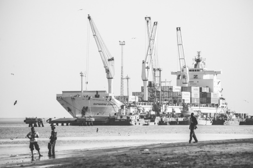 Banjul, The Gambia - February 21, 2011: Local people walking at the city beach, commercial port with a big container ship in background.