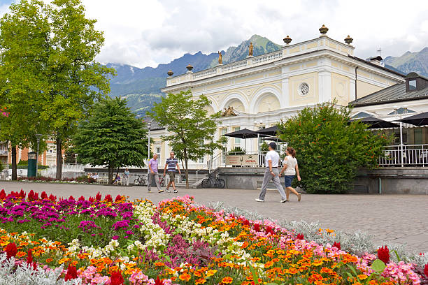 Merano, Italy - July 9, 2013: View to the Passer Promenade in Merano, South Tyrol, Italy. Tourists are visiting the town with the Mediterranean climate. In background is seen a part of the Kurhaus and the European Alps. The Kurhaus was built in 1912 and 1914 by the Viennese Jugendstil architect Friedrich Ohmann. In foreground is seen a colorful ornamental flower bed.