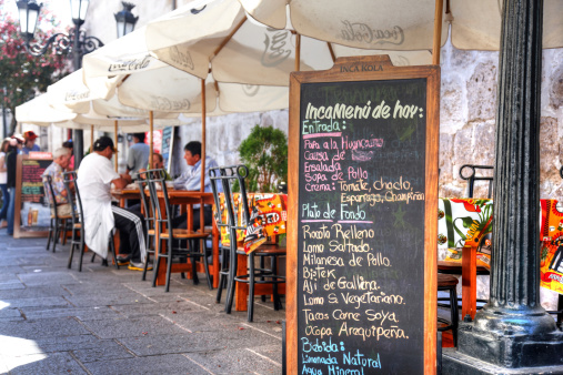 Arequipa, Peru - May 25, 2013: Restaurant menu on blackboard outside street cafes. Men and woman are pictured at the restaurant tables in sunlit cobblestoned street of Arequipa