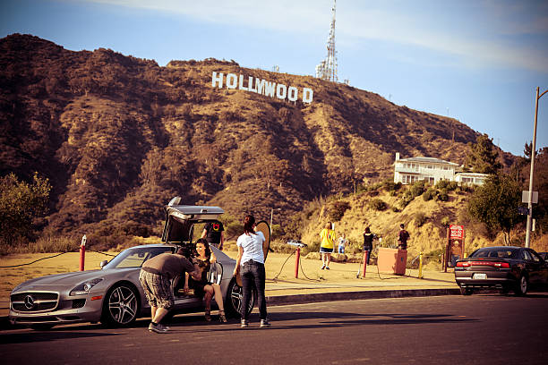 Hollywood, USA - May 3, 2013: Photographer shooting fashion model sitting in convertible Mercedes car with Hollywood Sign on background, assistant holding reflector