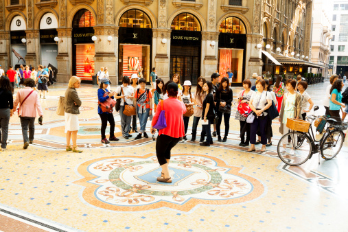 Milan, Italy - September, 1st 2013: A woman is turning foot on testicles of bull in mosaic inside of Galleria Vittorio Emanuele II. In background is standing a group of Asian women watching scene. At right side bicycle is standing. In background are shops.