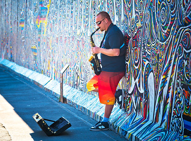 Berlin, Germany - September 7, 2013: Street musician playing the saxophone along the east side gallery wall. Graffiti paintings on the Berlin wall that are part of the East Side Gallery. Around 100 artists from all over the world painted in 1990 this section of the wall.
