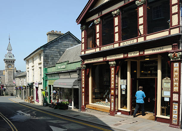 Hay-on-Wye, United Kingdom - July 12, 2013: Looking west down Lion Street, Hay-on-Wye, Powys, Wales, with one of the town's bookshops foreground. Also visible is a flower shop and an antique centre. In the distance is the clock tower, a Victorian Gothic design built in 1884., The town is famous for its bookshops although the number of them has declined rapidly in recent years, in part due to internet competition, and they have been replaced by women's clothing shops and antique shops or similar.Unidentified person entering shop.