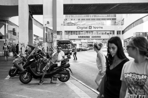 Tel Aviv, Israel - July 17, 2013: An Israeli man in his late 20s, listening to music and smoking a cigarette, sits on a motorbike parked at the Dizengoff Center on a hot summer day, watching other people come and go.