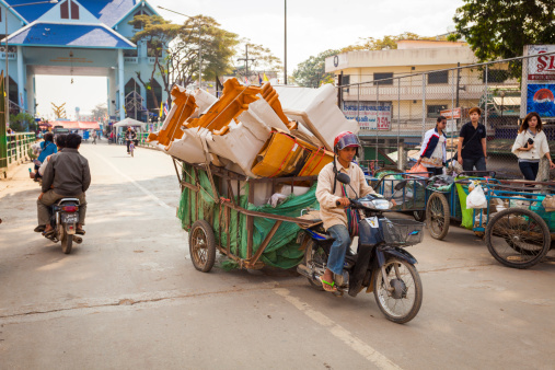 Mae Sai, Thailand - December 13, 2011: Motorcycle and rider loaded with goods on an attached trailer makes its way towards the border checkpoint at Mae Sai on the border between Thailand an Myanmar. The border area is busy with trade between the two countries. People can also be seen in the background.