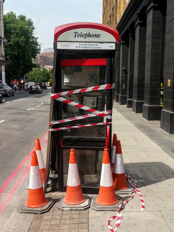 London, UK - July 16, 2013: An out of order BT telephone box, with cones and tape around it to prevent people from trying to use it, The telephone box is located on a pedestrian path besides a road, and you can see some cars in the background.
