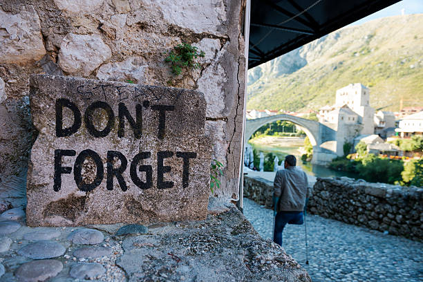 Don't Forget - Bosnian war in Mostar Mostar, Bosnia and Herzegovina - October 4, 2013: An amputee walks past a sign that reads, "Don't Forget", a reminder of the destructive war and seige that Mostar endured during the Bosnian war in the 1990s. In the background is the famous Old Bridge, or Stari Most, which was destroyed during the war and then rebuilt. stari most mostar stock pictures, royalty-free photos & images