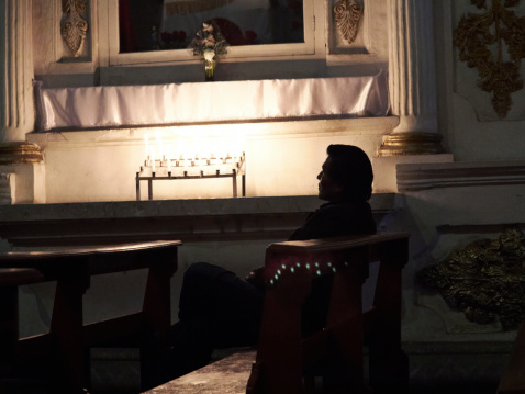 Puno, Peru - May 27, 2013: Mature hispanic man sitting in a pew praying in candlelit section of a Puno church