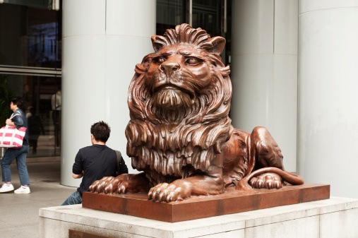 Hong Kong, China - March 31, 2010: One of the two HSBC guardian lions standing outside the bank in Hong Kong. This one is known as Stitt and the other as Stephen. A man with his back to us is sitting on the plinth at the back of the lion. In the background a woman is walking into the bank.