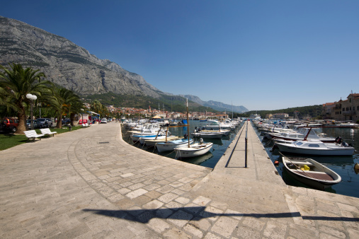 Makarska, A!roatia - August 25, 2011: Moored boats in the town of Makarska, few people in the distance walking enjoying the day, cars and houses also in the distance.