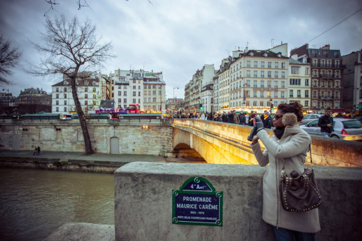 Paris, France - December 26, 2012: Young Woman Using Mobile Phone in Paris, standing on the bridge and photographing beautiful view on Seine River