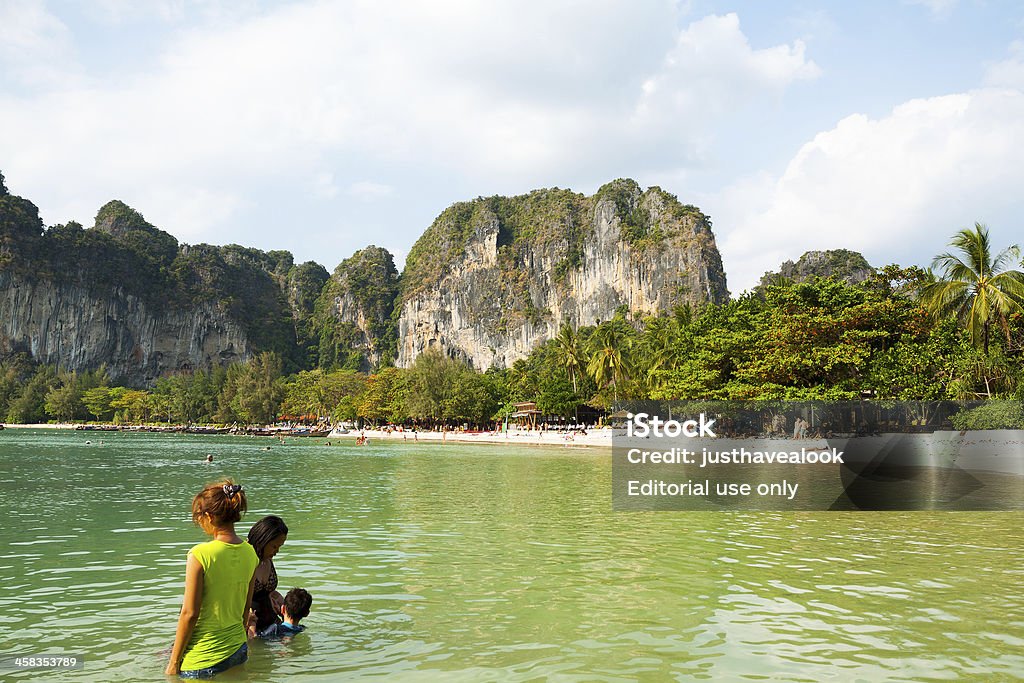 In sea and bay Ao Nang, Thailand - March, 4th 2013: View back from sea and bay to beach of small island offshore of Ao Nang. In water are some Thai women with child. On beach are many tourists around hotel areas. In background are rocks and tropical woods. Adult Stock Photo