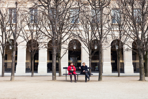Paris, France - March 5, 2013: two Parisian women rest on bench in Palais Royal garden in Paris in front of aracades of Palais Royal building. Originally called Palais-Cardinal, the palace was residence of Cardinal Richelieu and was built in 1639.