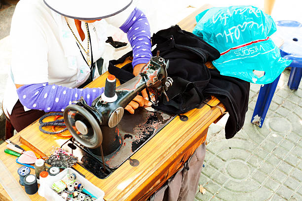 Bangkok, Thailand - February, 18th 2013: Female Thai dressmaker is at work. Woman is sitting with sewing machine in street in front of Tha Tien pier close to Wat Pho. She is wearing a hat against sun.